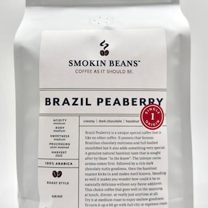 May include: A white stand-up pouch of Smokin Beans Brazil Peaberry coffee. The label features the brand name, product name, a single origin seal, and flavor notes of creamy, dark chocolate, and hazelnut. The net weight is 32 oz or 907 g.
