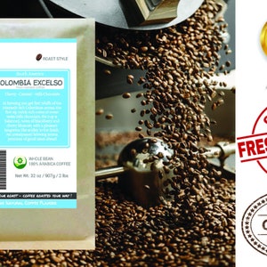 May include: A bag of whole bean Colombian Excelso coffee with a blue and white label that says "Smokin Beans Specialty Coffee Roastery". The label also says "Cherry - Caramel - Milk Chocolate" and "Whole Bean 100% Arabica Coffee". The bag is sitting on a pile of roasted coffee beans.