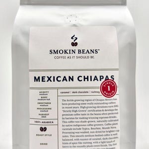 May include: A white coffee bag labeled Smokin Beans Mexican Chiapas. The label lists 100% Arabica, medium acidity, medium full body, and notes of caramel, dark chocolate, and nutmeg. The bag indicates a net weight of 32 oz, 2 pounds, or 907 g.