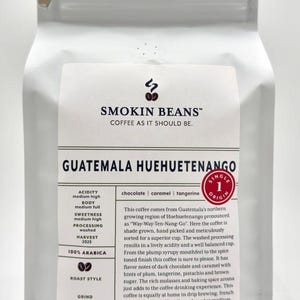 May include: A white coffee bag featuring the Smokin Beans logo and text. The label specifies Guatemala Huehuetenango, 100% Arabica, and lists flavor notes of chocolate, caramel, and tangerine. The net weight is 32 ounces or 2 pounds, 907 grams.