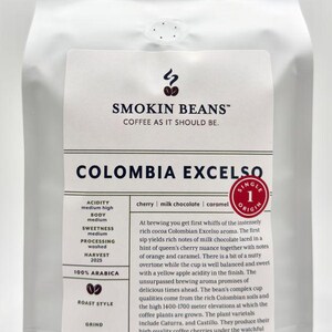 May include: A white coffee bag labeled Smokin Beans Colombia Excelso. The label lists 100% Arabica, medium acidity, medium body, and medium sweetness. It notes flavor notes of cherry, milk chocolate, and caramel. The bag weighs 32 oz or 2 pounds.