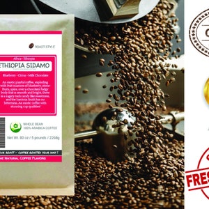 May include: A bag of whole bean Ethiopian Sidamo coffee with a pink and green label. The label features the text "Smokin Beans" and "Ethiopia Sidamo" with a description of the coffee's flavor profile: "Blueberry - Citrus - Milk Chocolate". The bag is sitting on a table with a coffee roaster in the background.