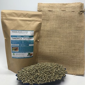 May include: A brown paper bag with a label that reads "Smokin Beans Coffee" and "Tanzania Peaberry" green coffee beans. The bag is sitting on a burlap sack. The coffee beans are in a blue bowl.