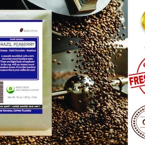May include: A brown paper bag with a white label that reads "Smokin Beans Specialty Coffee" and "Brazil Peaberry". The label describes the coffee as "Creamy - Dark Chocolate - Hazelnut" and notes that it is "Whole Bean 100% Arabica Coffee". The bag is sitting on a pile of roasted coffee beans.