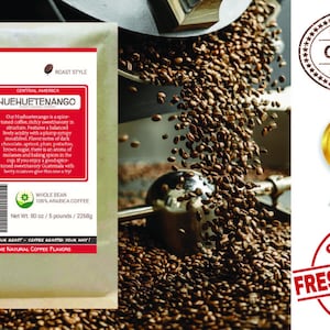 May include: A brown paper bag with a white label that reads "Smokin Beans Specialty Coffee Roasters" and "Huehuetenango" in bold text. The label also includes information about the coffee's origin, roast style, and weight. The bag is sitting on a pile of roasted coffee beans.