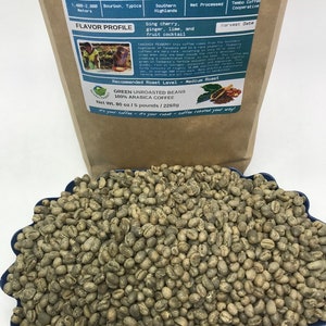 May include: A brown paper bag with the text "Tanzania Peaberry Green Coffee Beans" printed on it. The bag lists the elevation, varietal, region, process, farm, and harvest date of the coffee beans. The bag also includes a flavor profile and a recommended roast level. The bag is sitting on a blue surface with a pile of green coffee beans in front of it.