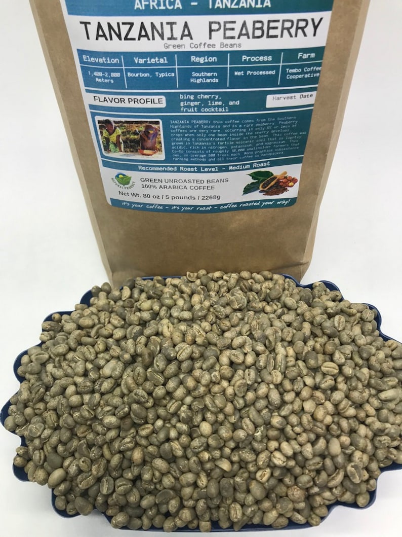 May include: A brown paper bag with the text "Tanzania Peaberry Green Coffee Beans" printed on it. The bag lists the elevation, varietal, region, process, farm, and harvest date of the coffee beans. The bag also includes a flavor profile and a recommended roast level. The bag is sitting on a blue surface with a pile of green coffee beans in front of it.