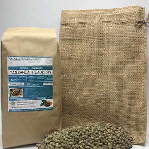 May include: A brown paper bag with the text "Smokin Beans Coffee" and "Tanzania Peaberry" on it. The bag contains green unroasted coffee beans. The beans are also shown in a blue bowl and a burlap sack.