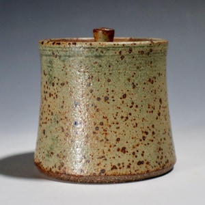 May include: A handmade ceramic jar with a lid. The jar has a cylindrical shape with a slightly flared base. It is speckled with brown spots and has a light green and tan color. The lid is small and brown.