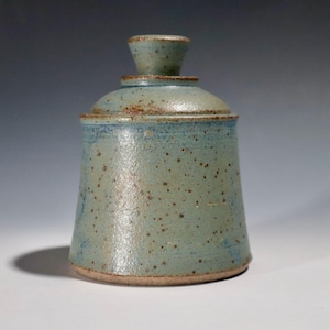 May include: A handmade ceramic jar with a lid. The jar is a muted teal color with brown speckles and a light blue rim. The lid has a small knob on top. The jar has a rustic, handcrafted appearance.