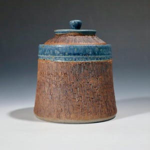 May include: A cylindrical ceramic jar with a textured, reddish-brown exterior featuring vertical grooves. The upper rim and the matching lid are finished in a smooth, glossy blue glaze. The lid has a small, central knob handle on top.