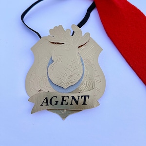 May include: A gold-colored badge with a deer head design and the word "AGENT" on a banner. The badge is attached to a black elastic band with a red felt bow tie.