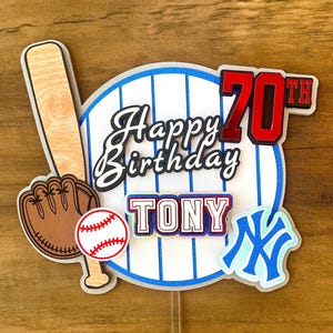 May include: A baseball-themed cake topper featuring a white circular background with blue pinstripes, a wooden bat, a brown glove, a white baseball with red stitching, a blue NY logo, and text reading Happy Birthday 70th TONY in red and black.