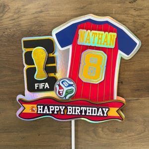 May include: A decorative cake topper featuring a soccer jersey with the name NATHAN and number 8, a FIFA World Cup trophy icon, a soccer ball, and a banner with the text HAPPY BIRTHDAY. The design includes red, yellow, blue, and black colors.