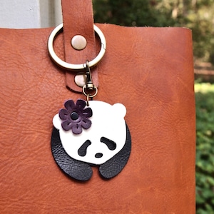 May include: A white and black leather panda keychain with a purple flower. The keychain is attached to a brown leather tote bag.