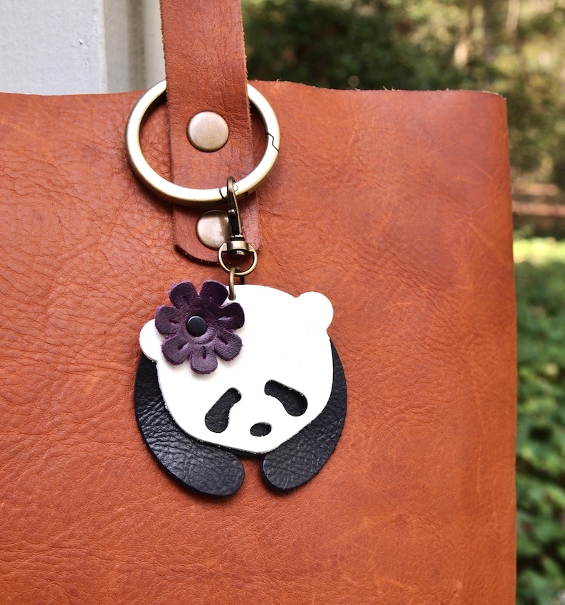 May include: A white and black leather panda keychain with a purple flower. The keychain is attached to a brown leather tote bag.