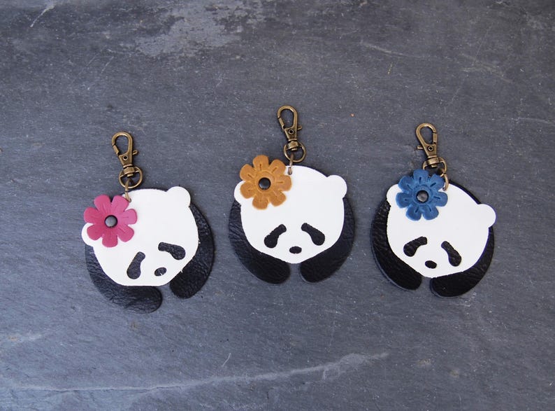 May include: Three leather panda keychains with different colored flower accents. The pandas are white with black ears and eyes, and the flowers are pink, yellow, and blue.