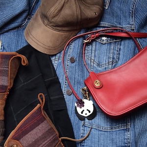 May include: A red leather shoulder bag with a brown leather strap and a panda charm. The bag is hanging on a blue denim jacket with a brown baseball cap.