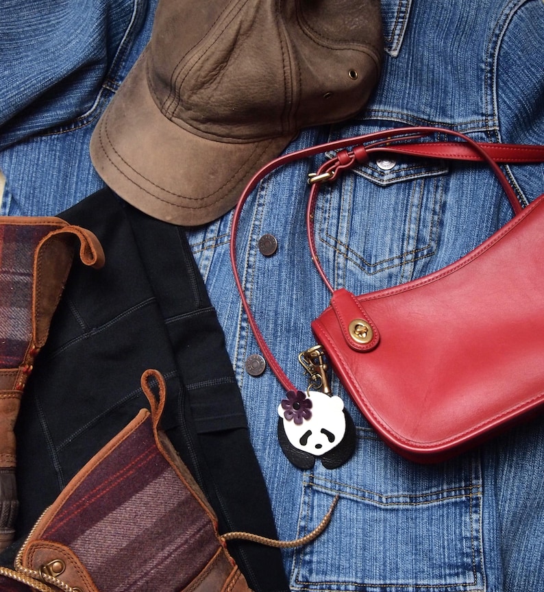 May include: A red leather shoulder bag with a brown leather strap and a panda charm. The bag is hanging on a blue denim jacket with a brown baseball cap.