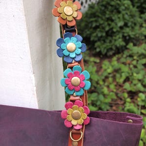 May include: A deep purple leather tote bag featuring a brown leather strap adorned with four layered flower embellishments. Each flower consists of two-toned leather petals in orange, blue, teal, pink, and yellow, centered with a brass-toned metal stud.