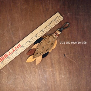 May include: A leather keychain with a brown, tan, and black leaf design. The keychain is about 4 inches long and has a metal clasp.