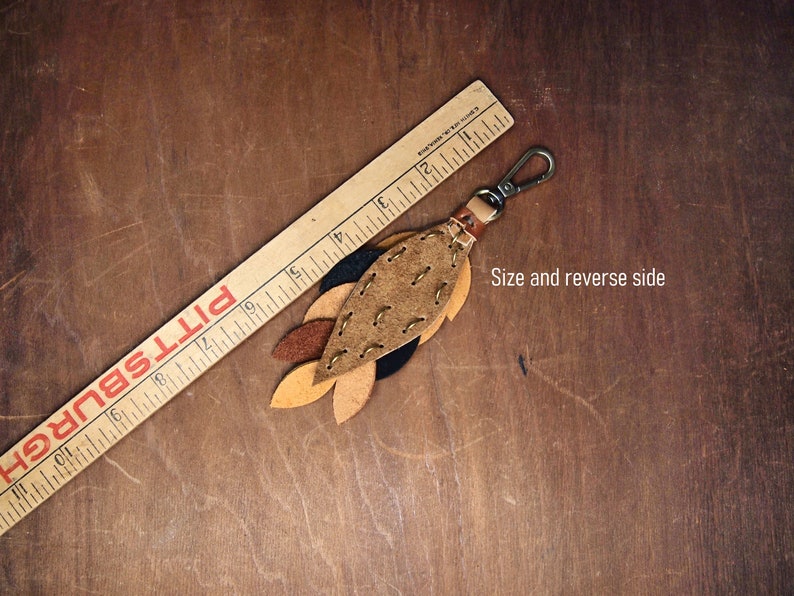 May include: A leather keychain with a brown, tan, and black leaf design. The keychain is about 4 inches long and has a metal clasp.