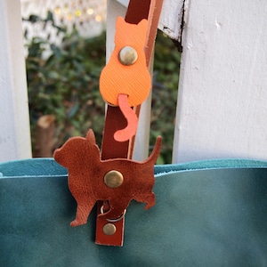 May include: A teal leather tote bag with a brown leather dog charm and an orange leather cat charm. The charms are attached to the bag's strap with metal rings.