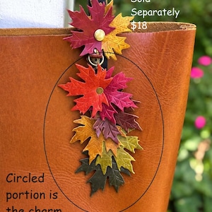 May include: A decorative bag charm featuring a cluster of layered maple leaf shapes in autumn colors including red, orange, yellow, and dark green. The leaves are attached to a central metal ring and hang from a dark strap. Text on the image reads: Flair Holder Sold Separately $18. Circled portion is the charm, flair holder at the top can be purchased separately.