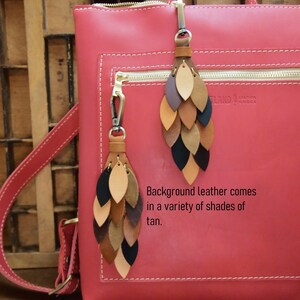 May include: A red leather backpack with two leather tassels. The tassels are made of multiple layers of leather in shades of brown, tan, and black. The backpack has a zipper closure and a gold zipper pull. The text "Leather Goods" is visible on the backpack.