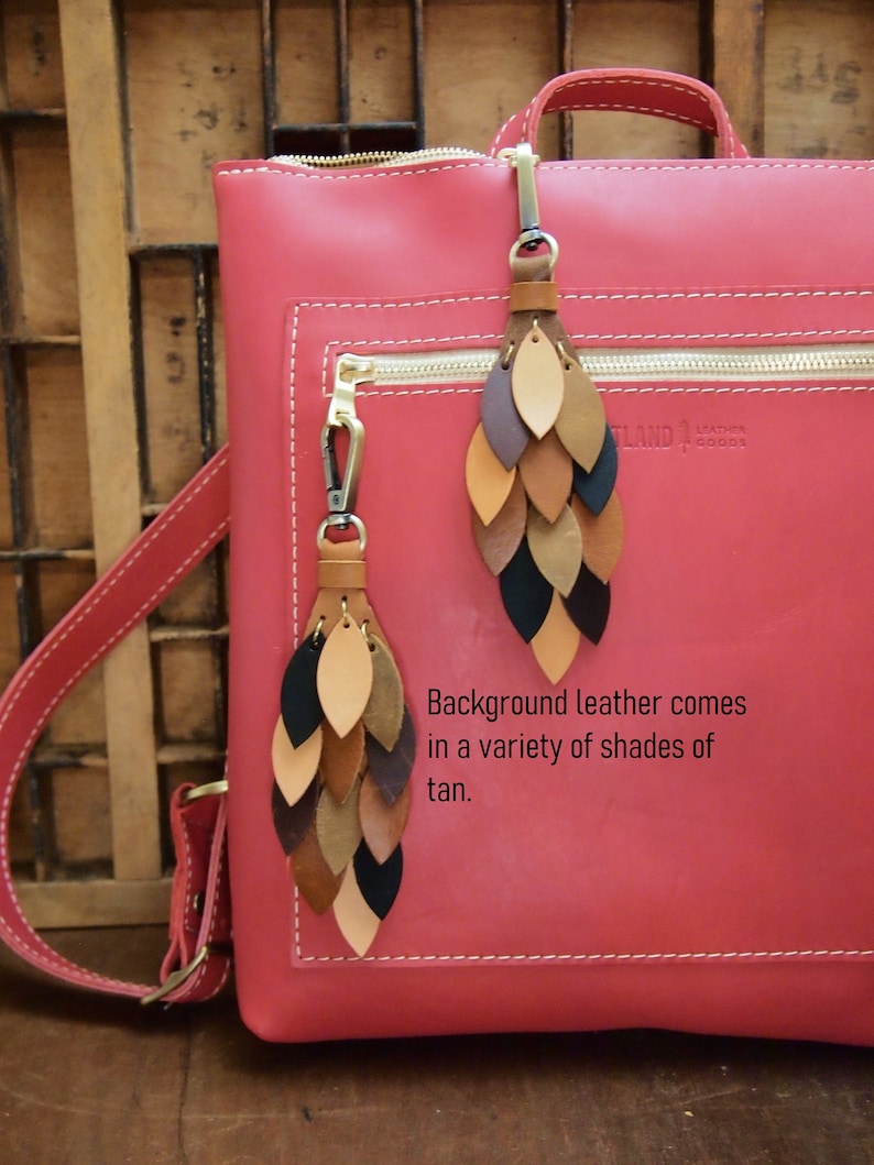 May include: A red leather backpack with two leather tassels. The tassels are made of multiple layers of leather in shades of brown, tan, and black. The backpack has a zipper closure and a gold zipper pull. The text "Leather Goods" is visible on the backpack.