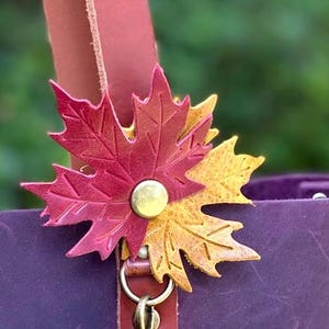 May include: A deep purple leather bag featuring a decorative charm shaped like two overlapping maple leaves. One leaf is a vibrant magenta and the other is a golden yellow. The leaves are secured with a central brass-toned metal rivet.