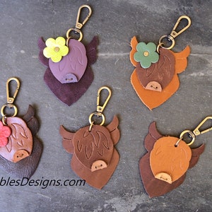 Op de afbeelding: Zes leren Highland Cow sleutelhangers met verschillende gekleurde bloemen. De sleutelhangers zijn bruin, paars en oranje. De bloemen zijn rood, groen, geel en paars.