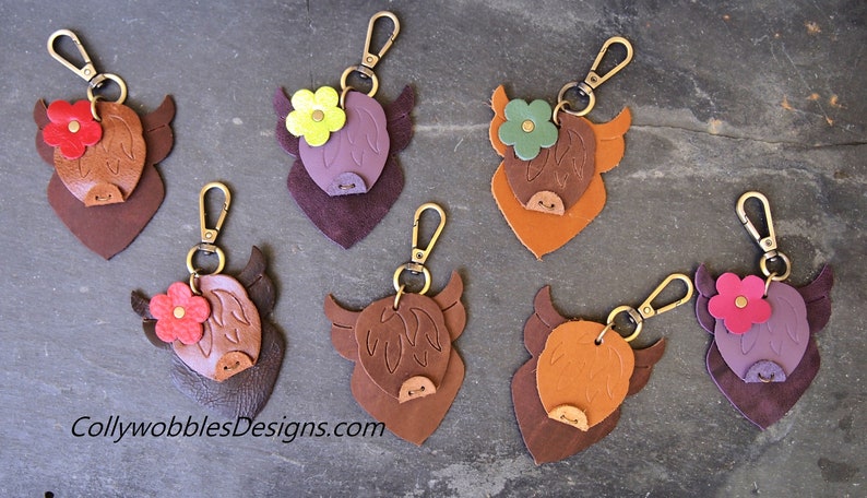 Op de afbeelding: Zes leren Highland Cow sleutelhangers met verschillende gekleurde bloemen. De sleutelhangers zijn bruin, paars en oranje. De bloemen zijn rood, groen, geel en paars.