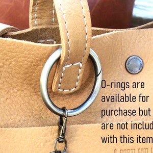 May include: Close-up of a tan leather tote bag with a silver O-ring attached to the handle. The bag has a white stitch detail and a small metal tag that reads "Portland Leather 1200". The text "O-rings are available for purchase but are not included with this item" is written below the bag.