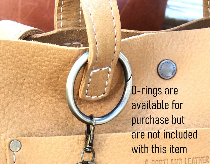 May include: Close-up of a tan leather tote bag with a silver O-ring attached to the handle. The bag has a white stitch detail and a small metal tag that reads "Portland Leather 1200". The text "O-rings are available for purchase but are not included with this item" is written below the bag.