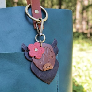 Op de afbeelding: Een teal leren tote bag met een bruine leren band en een sleutelhanger met een bruin en zwart leren hoogland rund met een rode bloem.
