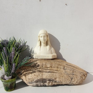 Könnte beinhalten: Weiße Gipsbüste einer Frau mit weißem Kopftuch, die auf einem Stück Treibholz sitzt, mit einer kleinen Topfpflanze mit lila Blüten im Vordergrund.