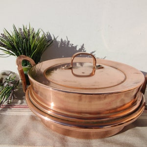 以下が含まれることがあります： 2つのハンドルと蓋が付いたヴィンテージの銅製の蒸し器。蒸し器はストライプの布の表面の上に置かれています