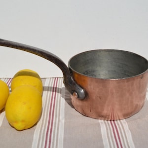 May include: A round copper saucepan with a hammered exterior finish and a long, dark, riveted metal handle. The interior of the pan is silver-toned metal. Three bright yellow lemons are positioned to the left of the copper cookware piece.