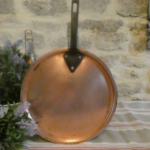 May include: A copper pan with a black handle. The pan is round and has a shiny copper finish. The handle is attached to the pan with three rivets.