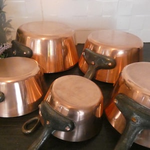 May include: A collection of copper pots with dark handles. The pots are arranged in a group, showcasing their shiny, reflective surfaces. The handles are a dark, possibly cast iron, material. The pots vary in size and shape.