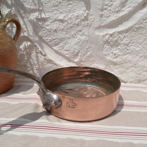 May include: A round copper saut&eacute; pan with a long, dark metal handle attached by three rivets. The exterior of the pan features a stamped mark with the number 20 and the text Made in France. The interior of the pan has a metallic lining.