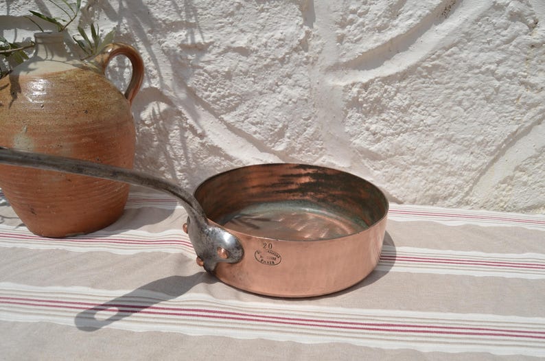May include: A round copper saut&eacute; pan with a long, dark metal handle attached by three rivets. The exterior of the pan features a stamped mark with the number 20 and the text Made in France. The interior of the pan has a metallic lining.