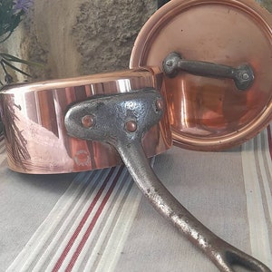 Puede incluir: Una cacerola de cobre con tapa y un mango de hierro negro. La cacerola tiene un acabado martillado y el mango tiene remaches.