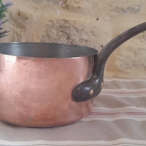 Op de afbeelding: Een koperen pan met een houten handvat. De pan staat op een gestreept tafelkleed.