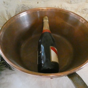 May include: A copper pot with a bottle of champagne inside. The pot has a handle and is sitting on a white tablecloth.