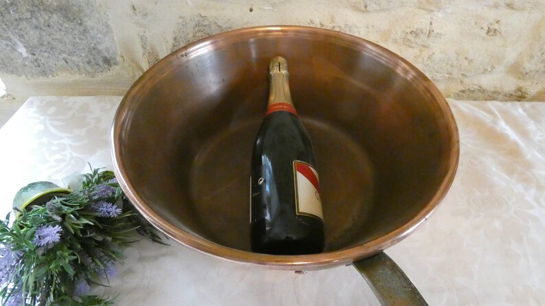 May include: A copper pot with a bottle of champagne inside. The pot has a handle and is sitting on a white tablecloth.