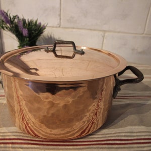 May include: A large copper pot with a hammered finish and a lid. The pot has two black iron handles and a single black iron handle on the lid.