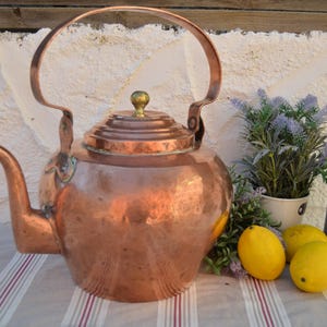 Puede incluir: Una gran tetera de cobre antigua con asa y boquilla curvadas. La tetera se exhibe con una botella de champán, limones y una pequeña planta en maceta. La tetera de cobre tiene una superficie brillante y reflectante.