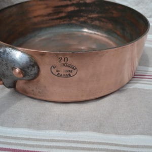 May include: A round copper saut&eacute; pan with a dark metal handle attached by three copper rivets. The side of the pan features the stamped text 20 and an oval mark reading Grands Magasins du Louvre Paris. The interior shows a metallic finish.